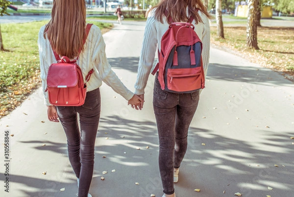 Fototapeta Two teenage girls walk in city on autumn day in summer, hold hands, look from back, casual clothes, jeans and a sweater, walk after school and college. Backpacks with textbooks and abstracts behind.