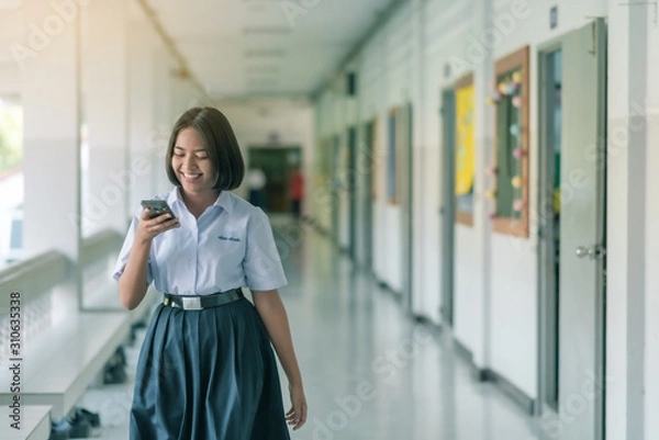 Fototapeta A smiling Asian female high school student in white uniform is walking and enjoying social media on her smartphone in the school.