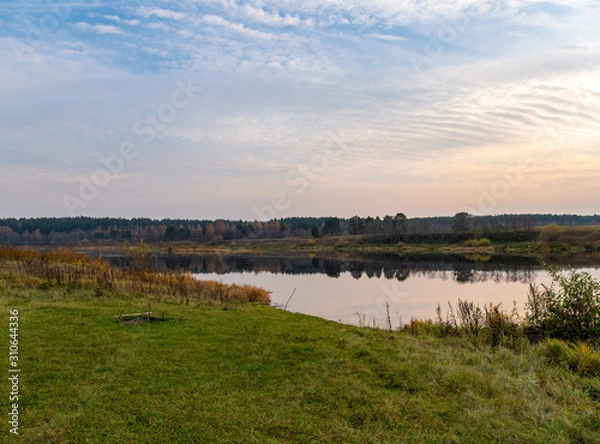 Fototapeta Calm landscape at eve dusk. Green bank with a place for relaxation and a picnic with beautiful view of the river, distant forest. The still mirror of the water reflects trees and sunset in cloudy sky
