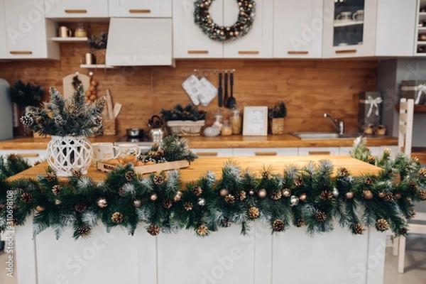 Fototapeta View over beautiful white kitchen with Christmas decorations all over cupboards and kitchen board. There is Christmas wreath on the cupboard. Natural fir tree branches with pine cones.