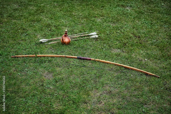 Fototapeta Ancient ceramic jug, wooden long bow and arrows lying on the ground with green grass. Historical reenactment concept, hobbies and leisure