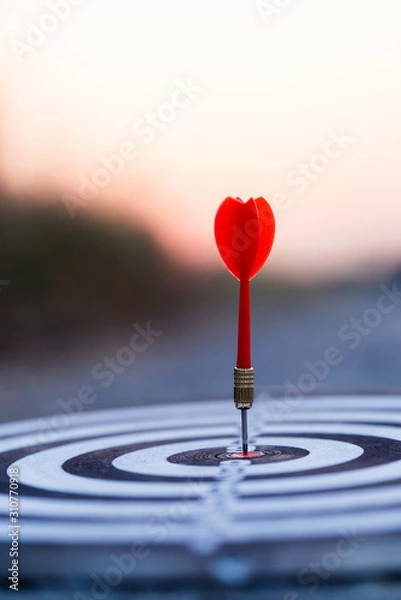 Obraz Close up red dart arrow hitting target center dartboard on sunset background