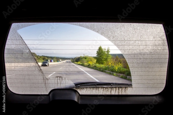 Obraz dirty car view from inside the car through the rear window, rear window of a dirty car with a view of the asphalt road with markings and road traffic.