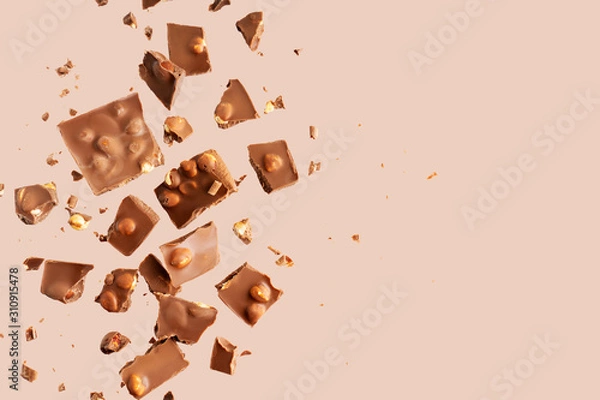 Fototapeta Flying in the air broken bar of milk chocolate with nuts and flakes on pastel pink background.  Chocolate pieces levitation concept.