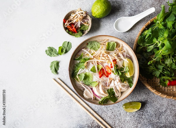 Fototapeta Traditional Vietnamese soup- pho ga in bowl with chicken and rice noodles, mint and cilantro, red onion, chili, bean sprouts and lime on grey background. Asian food. Copy space. Top view
