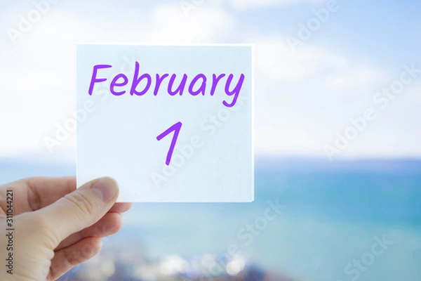Obraz February 1st. Hand holding sticker with text February 1 on the blurred background of the sea and sky. Copy space for text. Month in calendar concept