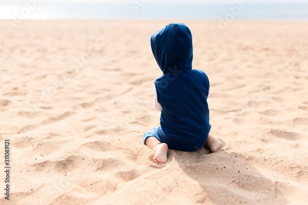 Fototapeta  unrecognizable child sits with his back in a sweater with a hood on the beach with bare legs.