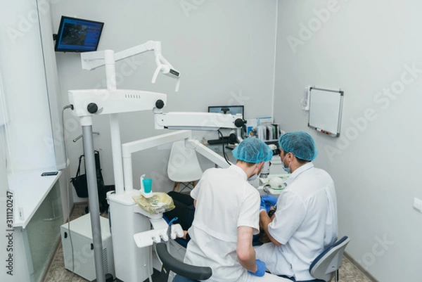 Fototapeta Doctor used microscope. Dentist is treating patient in modern dental office. Operation is carried out using cofferdam. Client is inserted and restored teeth, make denture. Orthodontist and assistant
