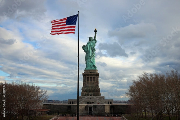 Fototapeta Statue of Liberty from behind with waving flag of the United States of America in autumn and cloudy weather