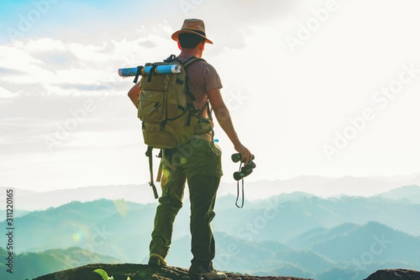 Fototapeta The hiker with a backpack stood on the rock after examining the map to find a path in a beautiful mountain landscape.