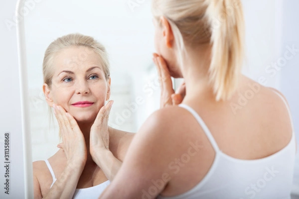 Obraz Middle aged woman looking at wrinkles in mirror. Plastic surgery and collagen injections. Makeup. Macro face. Selective focus