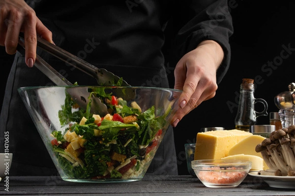 Fototapeta Cooking salad, mixing in a large transparent plate. On a black background with ingredients. Cooking, gastronomy, home recipes. Recipe book, delicious food concept. Home cooking, Greek or Italian salad
