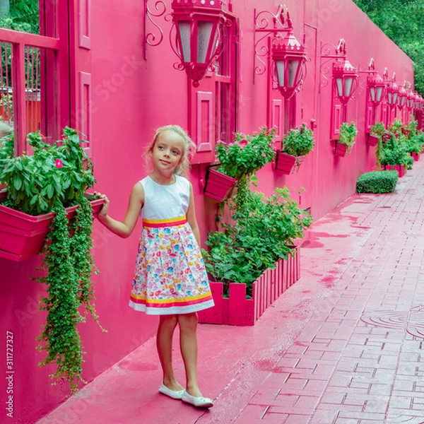 Obraz Little girl walking in a pink street with green plants, windows with shutters, lamps, decorative caribbean entourage in old city victorian style, Puerto plata, Dominican Republic, Paseo de doña Blanca