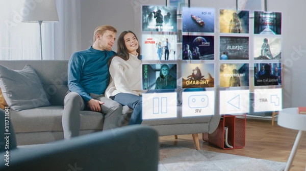 Obraz Beautiful Young Couple are Sitting on a Couch at Home and Choosing a Movie to Watch on a Futuristic Hologram Screen.