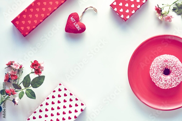 Fototapeta Valentine day flat lay, top view on white table. Geometric background with pussy willow. Gift boxes, pink doughnut on red plate and heart in hand. German text "Mit liebe" on heart means "with love".