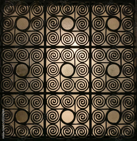 Fototapeta Wrought Greek iron gate grille, ornamented with elaborately twisted spirals and with frosted glass in the background, that is slightly illuminated. Metal sheet and barrier. Antique reproduction. Photo