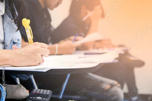 Fototapeta Hands university student holding pen writing /calculator doing examination / study or quiz, test from teacher or in large lecture room, students in uniform attending exam classroom educational school.