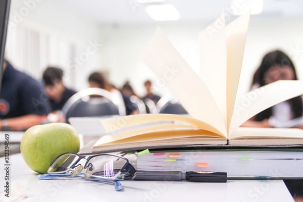 Fototapeta A notebook/pen/books/grasses of teacher on table and students during study or quiz, test and exam in large lecture room / University classroom. Students are in uniform classroom educational school.