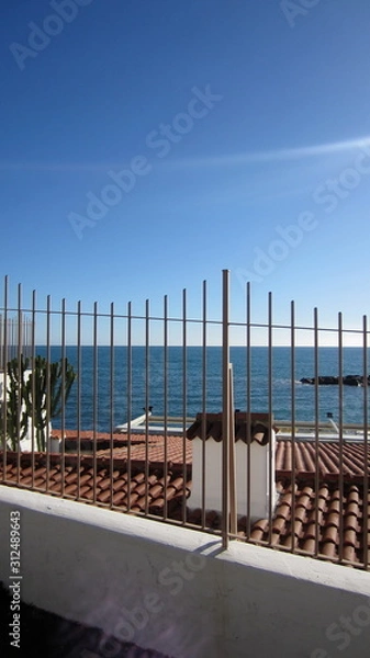 Fototapeta Metal fence of the house by the sea with a view of the roof, cactus and the sea on a sunny cloudless day