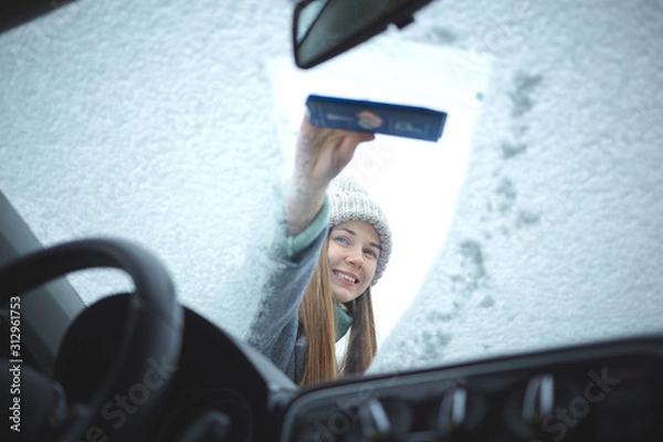 Fototapeta Smiling girl cleaning frozen windshield of a car from snow.