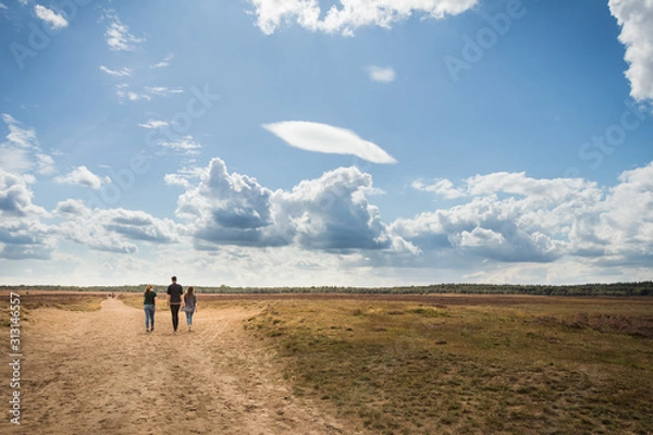 Obraz Three young people walking on a heathland, enjoying a beautiful blue sky with clouds, the path bends to the left and to the right.
