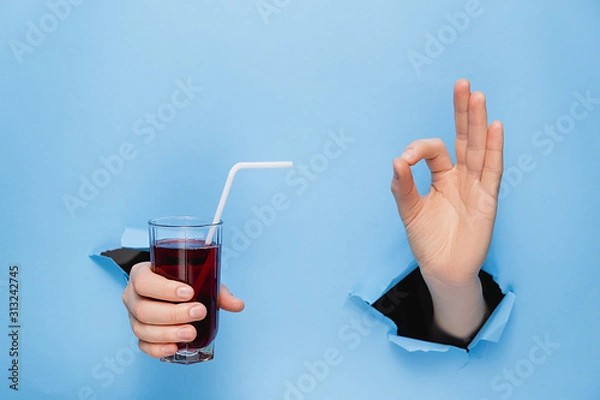 Fototapeta Close up of female hand holding a glass of fresh red juice with a straw through torn blue paper wall. Fruit drinks and healthy nutrition. Hand sign. Hole in wall. Like gesture