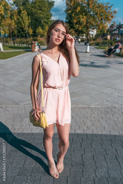 Obraz Beautiful girl in pink dress, stands in summer in city, adjusts glasses, yellow handbag, posing. Background road trees paving stones. Emotions of relaxation comfort on bright sunny day.