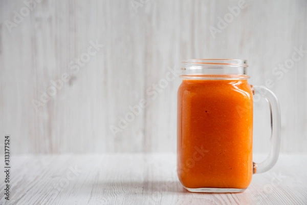 Fototapeta Homemade Mango Carrot Smoothie in a glass jar mug over white wooden background, side view. Copy space.