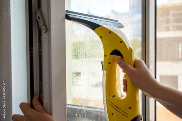 Obraz Wash the window at home with a battery wiper. The apparatus yellow collects drops of water and clean the window. Hands scrub the window. White window frame with a handle.