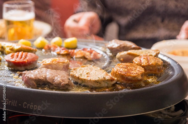 Fototapeta pieces of meat are roasting on a gourmet set. Smoke is coming from the hot plate. Beer and hand of person in background.