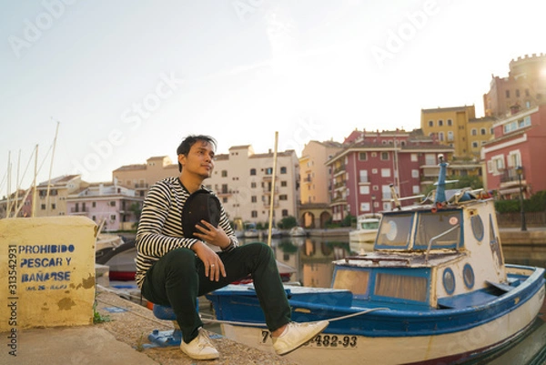 Fototapeta Asean young man taking a photo with black hat and smiling on the edge of a city river with boats and views of colorful houses in Valencia, Spain