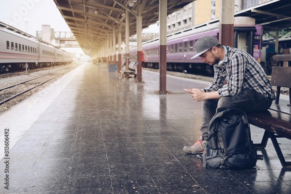 Fototapeta Young man using mobile smart phone checking train timetable at train station platform. Asian traveler text message with cellphone while waiting for train. Technology travel lifestyle concept.