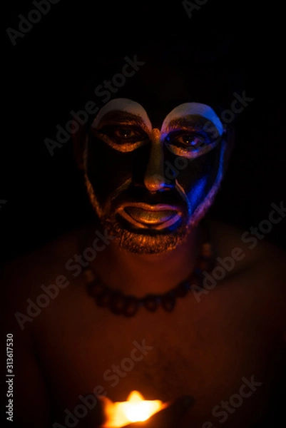 Obraz Close up of Indian brunette man with his face illuminated and painted by black and golden like a tribe standing with candle light in front of a black studio copy space background. Indian hi fashion.