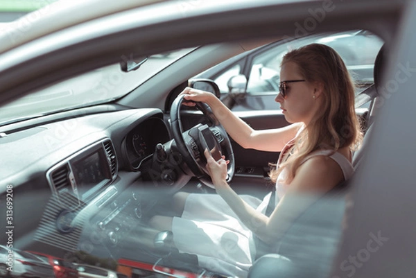 Fototapeta woman in car, sitting behind wheel of a car, reads and writes message in mobile phone, parking mall, traffic jam, in summer in car. Girl in pink dress, automatic transmission.