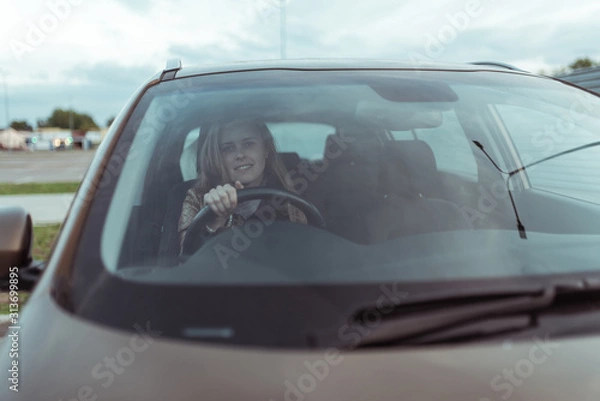 Fototapeta woman in car, sitting behind the wheel of a car, happy smile, right wheel of car, left-hand traffic. Windshield, fall day outside. Free space for copy text.