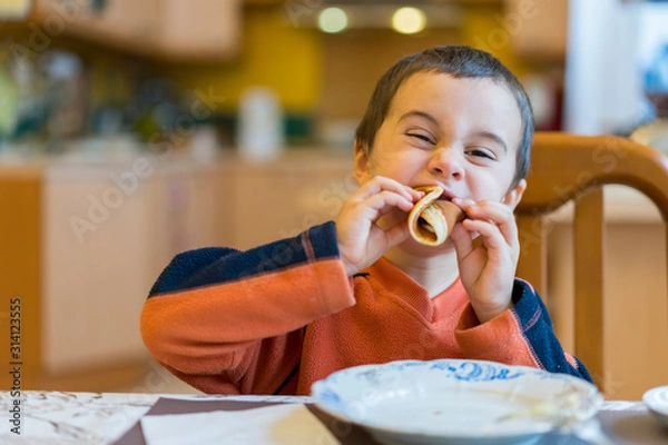 Fototapeta Happy little boy eats pancakes hands. Portrait of small cute little boy child caucasian sitting by the table at home eating pancake