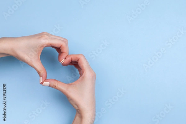 Fototapeta Female hands show a heart symbol on a light blue background. Place for text.