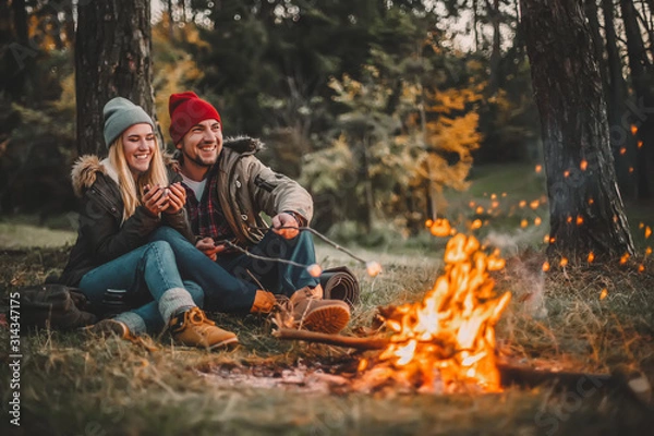 Fototapeta Traveler couple camping and roasting marshmallows over the fire in the forest after a hard day. Concept of trekking, adventure and seasonal vacation.