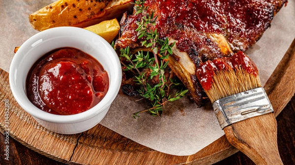 Obraz American cuisine. Fried pork and ribs in a marinade with homemade barbecue sauce, next to it lies a marinade brush. Serve on a wooden kitchen board. background image, copy space