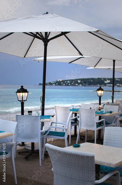 Obraz Tables for a romantic meal on the beach with lanterns and chairs. With sunset view. Menton, France
