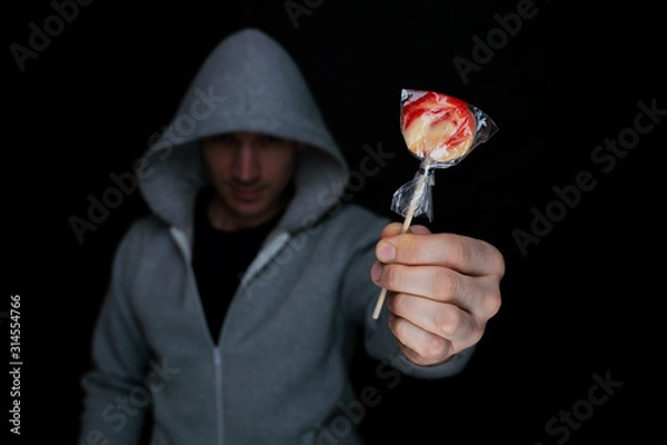 Fototapeta Angry man in a hood holds a lollipop in his hand and holding out to his victim, for luring. Selective focus