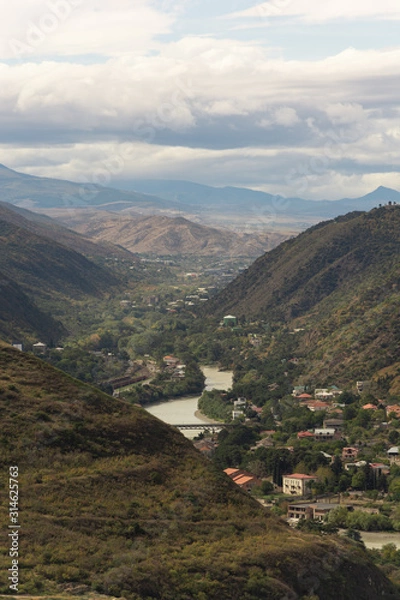 Obraz A magnificent view a valley surrounded by picturesque mountains, with numerous villages located along the river, over background of misty horizon and cloudy sky.