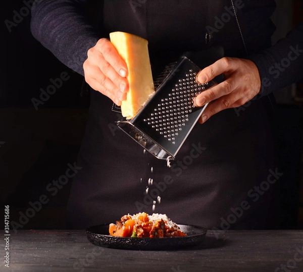 Fototapeta A man in an apron rubs cheese on a grater, prepares a dish. Cheese falls on food. Dark background.