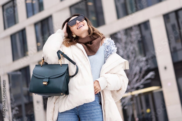Fototapeta European model in a beige oversized down jacket, knitted sweater with a handbag and glasses posing near the office building. Lifestyle. Real emotions
