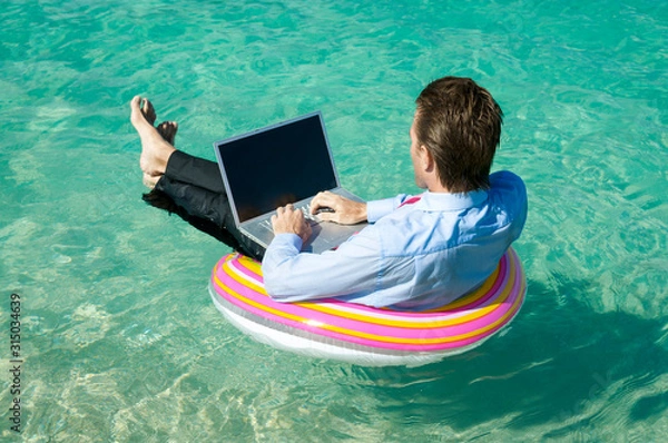 Fototapeta Relaxed businessman floating on a rubber ring while typing on his laptop