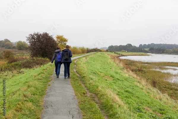 Fototapeta 2 female friends enjoying a walk along a river path on a dull and rainy autumn day
