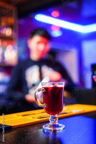 Fototapeta A transparent glass with mulled wine stands on a table on a blue background, behind a man stands on a blurred background, bright colors, side view