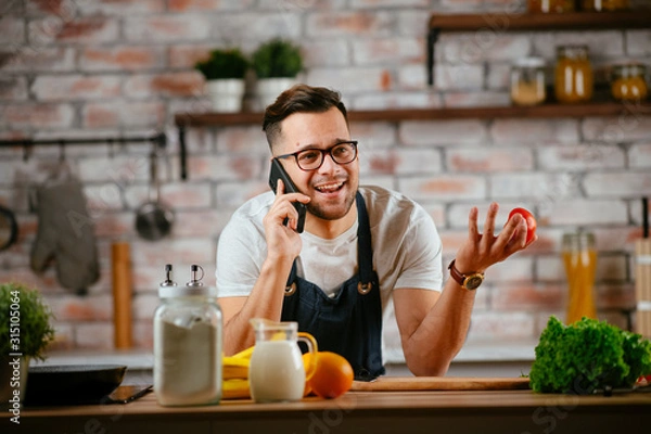 Fototapeta Young man in kitchen. Chef using phone while making food. 