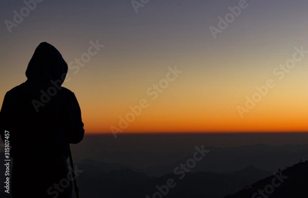 Fototapeta silhouette of Travel photographer standing with a camera mounted on a tripod and shooting a time lapse of the sunrise/sunset. man wearing his hood enjoying the mountain view /valley view