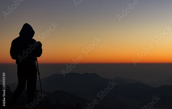 Fototapeta silhouette of Travel photographer standing with a camera mounted on a tripod and shooting a time lapse of the sunrise/sunset. man wearing his hood enjoying the mountain view /valley view